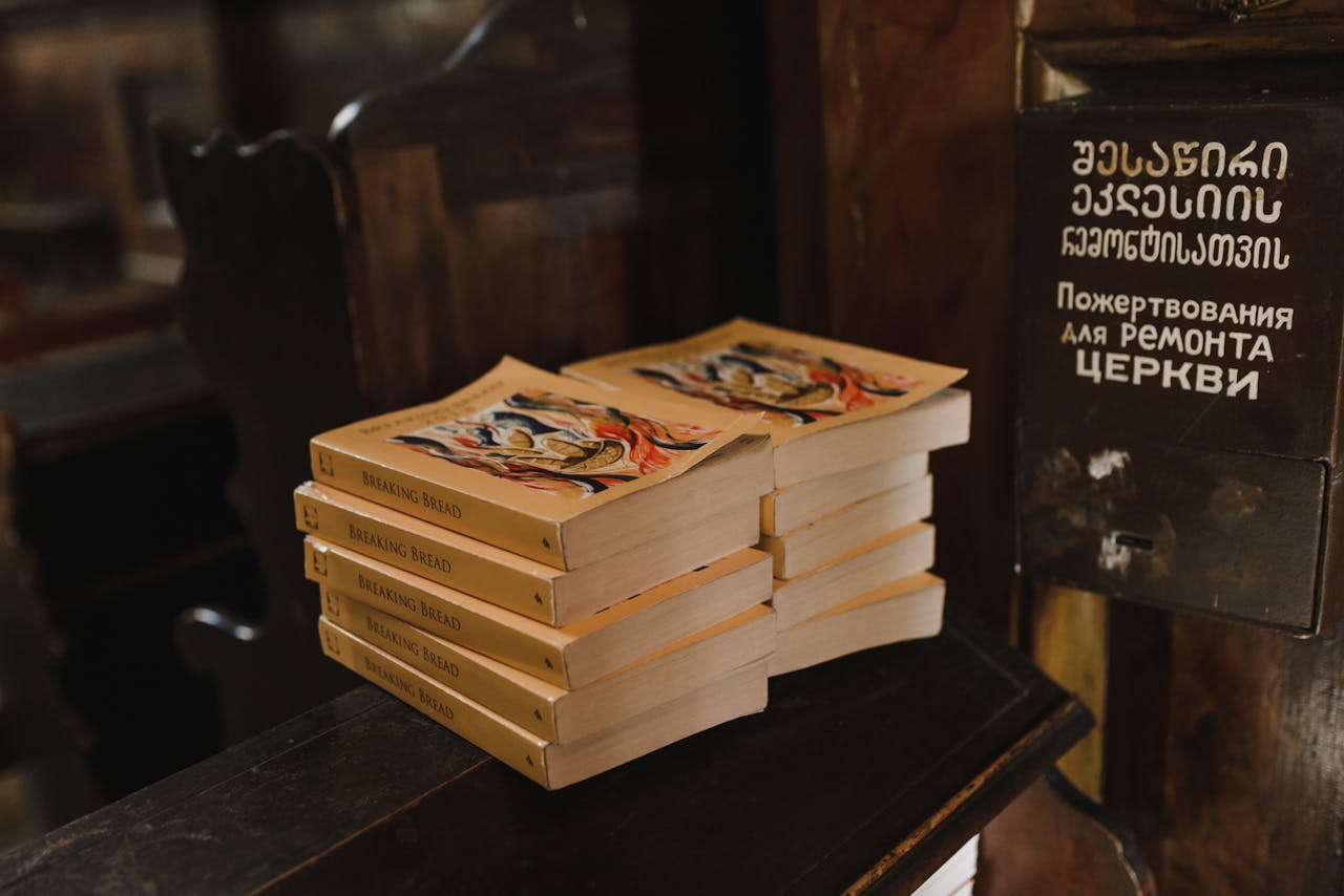 A stack of books titled 'Breaking Bread' on a wooden church surface with donation box.